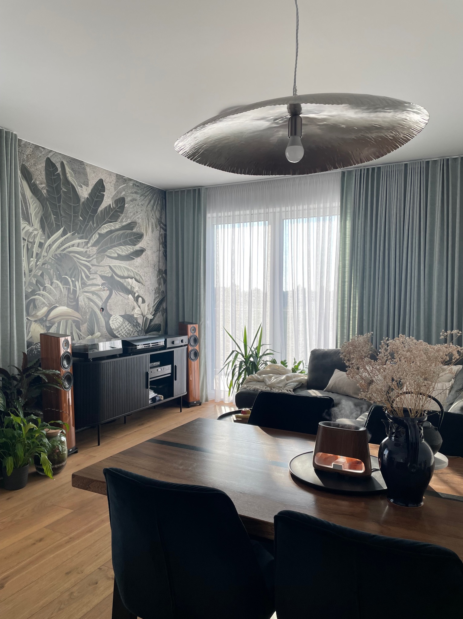 Salon z tapetą w tropikalne liście, drewnianym stołem i lampą w kształcie talerza. Widoczne głośniki i szafka RTV. Stonowana kolorystyka, naturalne światło.
