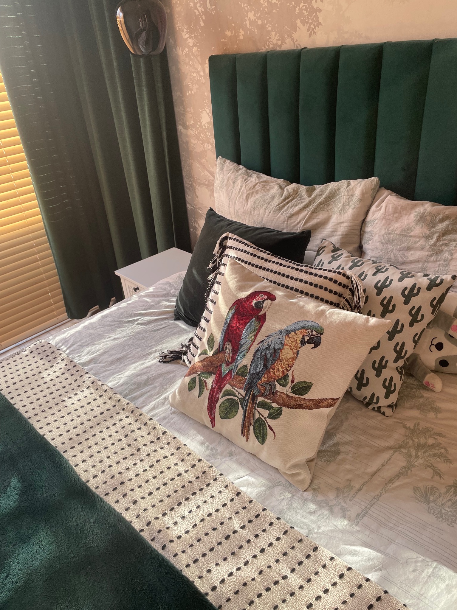 Przytulna sypialnia z zielonym zagłówkiem, dekoracyjnymi poduszkami z motywem papug i kaktusów, oraz zasłonami w odcieniach zieleni. Delikatne światło słoneczne wpada przez żaluzje.