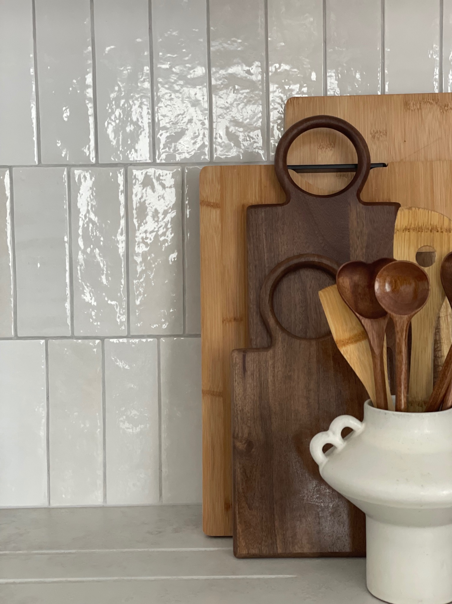 Białe, błyszczące płytki na ścianie kuchni, drewniane deski do krojenia i biały wazon z drewnianymi łyżkami. Minimalistyczny wystrój kuchni.