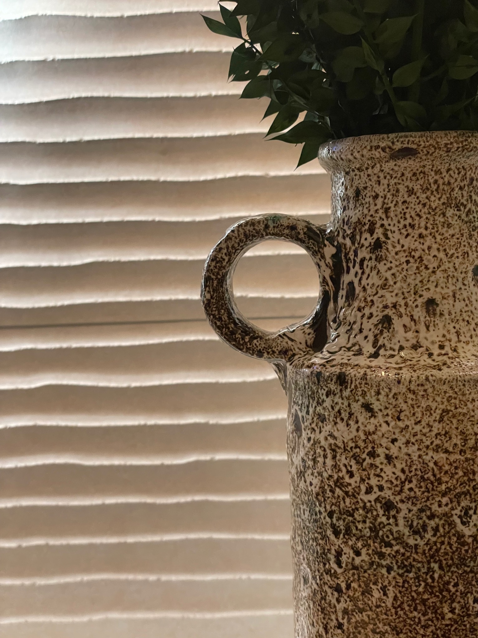 Dekoracyjny, ceramiczny wazon z uchwytem, wypełniony zielonymi gałązkami, na tle poziomych żaluzji. Neutralna kolorystyka i faktura wazonu.