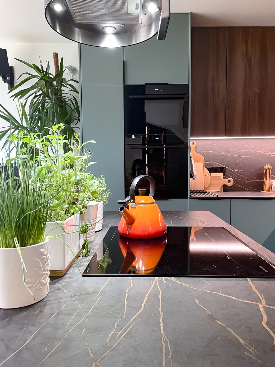 Nowoczesna kuchnia z marmurowym blatem, ziołami w doniczkach i pomarańczowym czajnikiem na płycie indukcyjnej. Minimalistyczne szafki w odcieniach szarości i brązu.