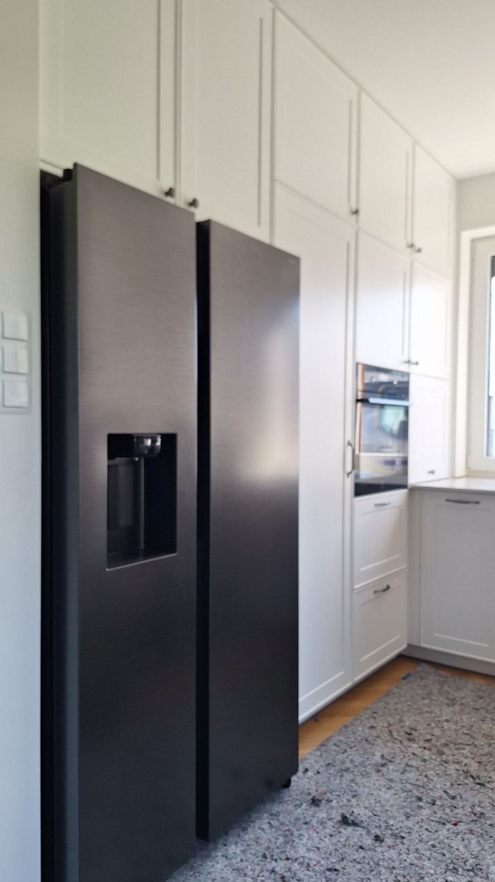 Nowoczesna kuchnia z zabudową meblową pod sufit i dużą, dwudrzwiową lodówką w kolorze grafitowym. Białe fronty szafek, piekarnik w zabudowie, szary dywanik.