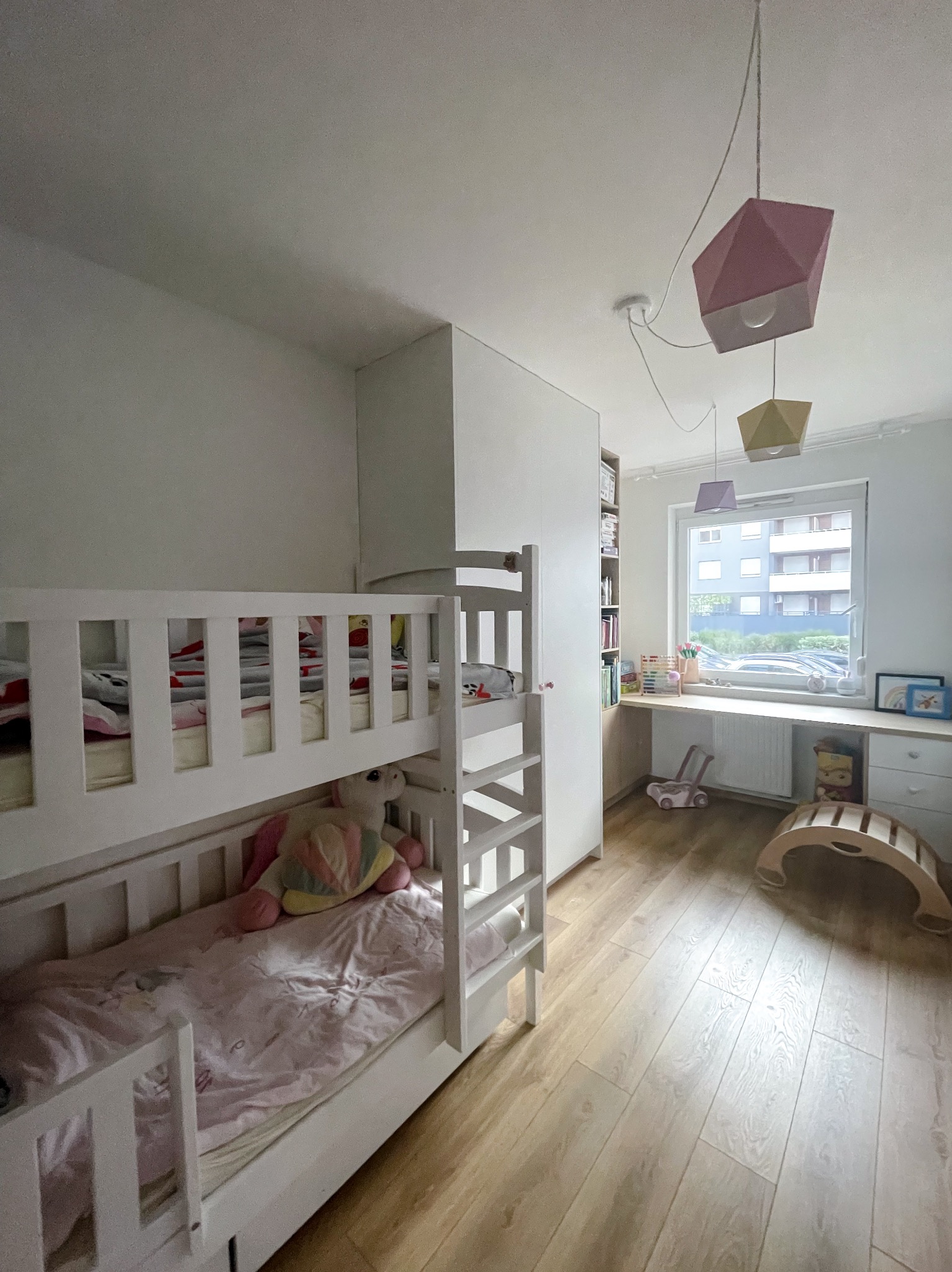 Pokój dziecięcy z białym łóżkiem piętrowym, biurkiem pod oknem i geometrycznymi lampami wiszącymi. Jasne drewniane panele podłogowe. Minimalistyczny styl.