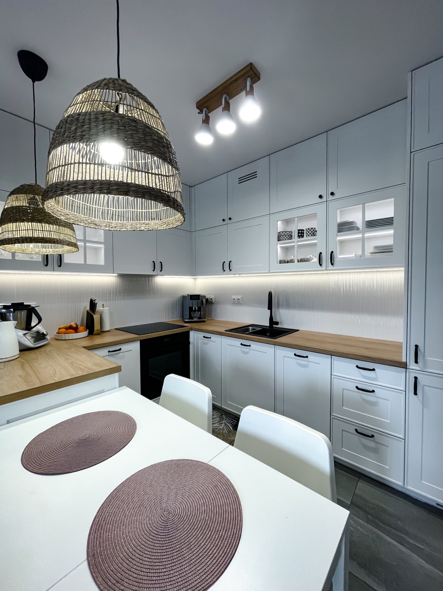 Jasna kuchnia z drewnianym blatem i wiklinowymi lampami. Białe szafki z czarnymi uchwytami. Na blacie ekspres do kawy i owoce. Minimalistyczny design.