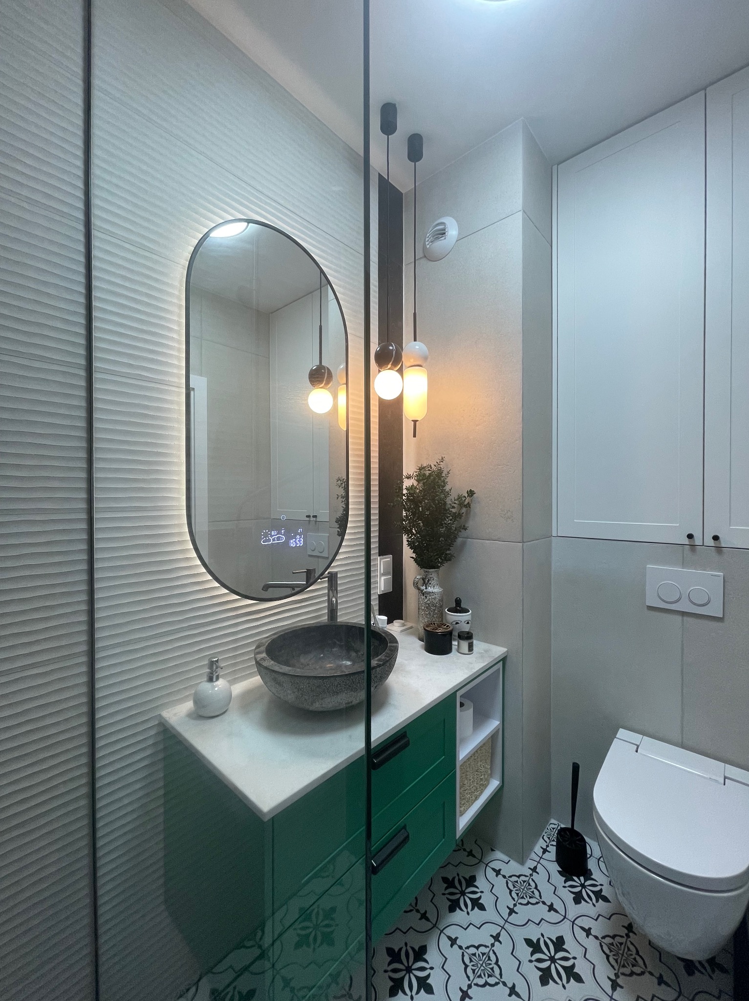 Nowoczesna łazienka z umywalką z kamienia, zieloną szafką i podświetlanym lustrem. W tle biała szafka i toaleta. Na podłodze wzorzyste płytki.