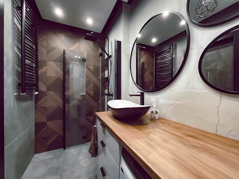 Nowoczesna łazienka z kabiną prysznicową, umywalką na drewnianym blacie i okrągłymi lustrami. Geometryczne płytki na ścianie dodają charakteru.