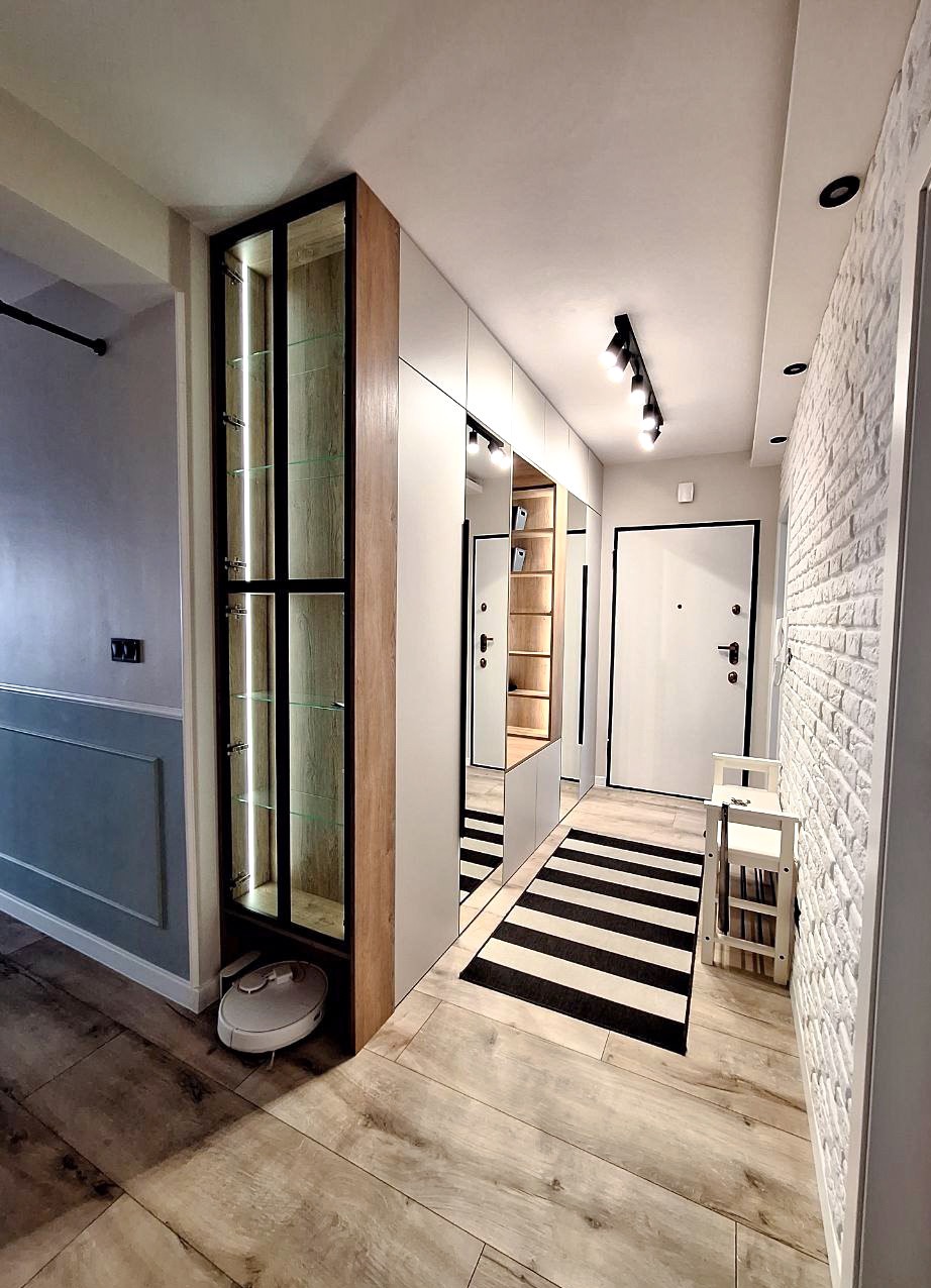 Nowoczesny przedpokój z szafą w zabudowie, lustrami i oświetleniem punktowym. Na podłodze dywan w czarno-białe pasy i drewnopodobne panele. Widoczny robot sprzątający.