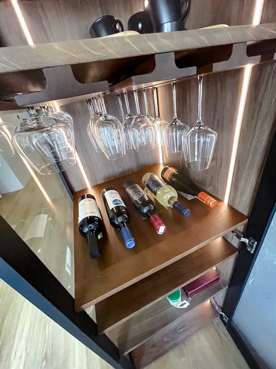 Drewniana szafka barowa z oświetleniem LED, eksponująca butelki wina i kieliszki. Nowoczesny design z ciemnymi akcentami i ciepłym światłem.