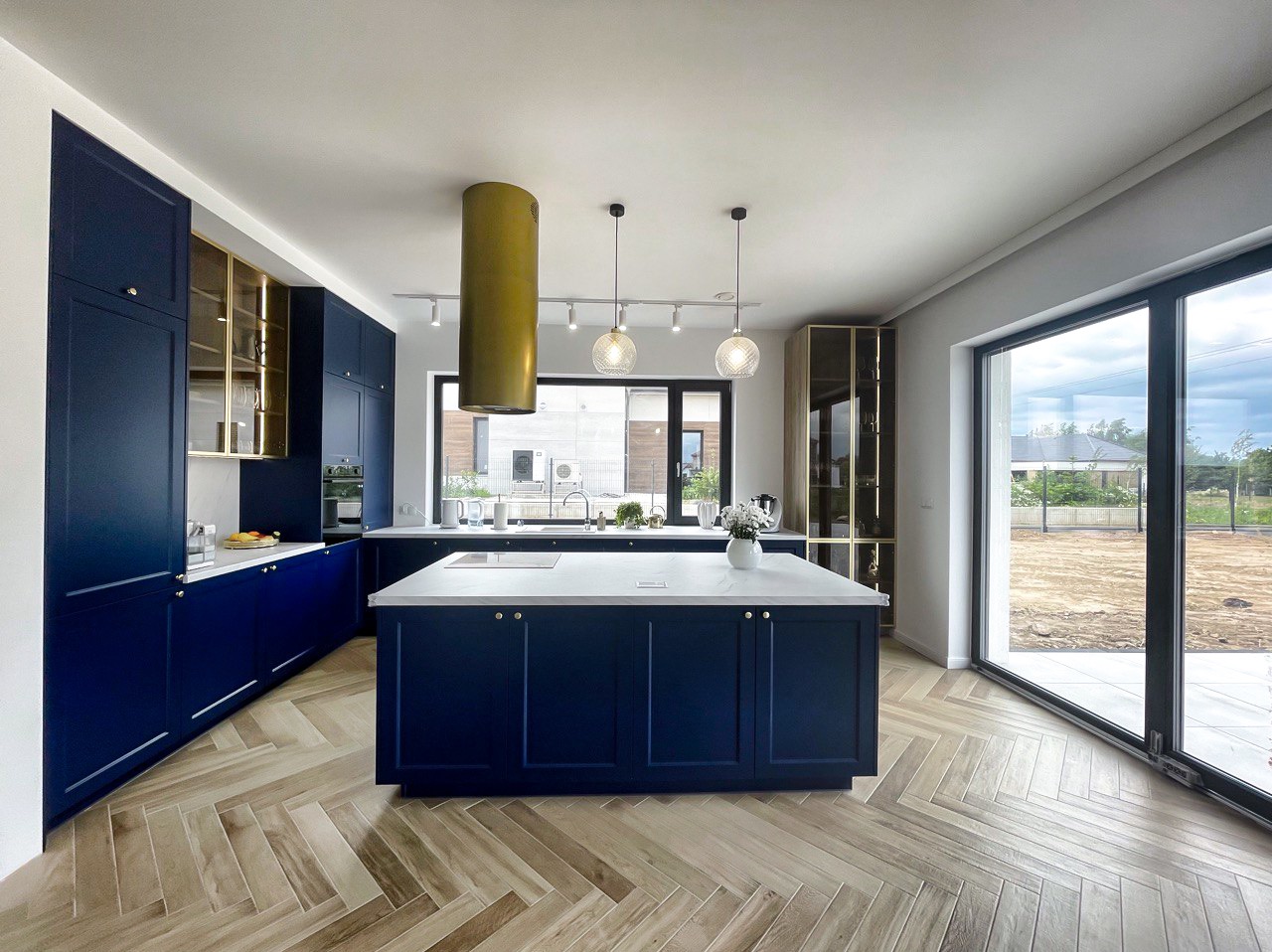 Nowoczesna kuchnia w odcieniach granatu z wyspą i złotymi akcentami. Parkiet ułożony w jodełkę, duże okno z widokiem na zewnątrz. Minimalistyczny design.