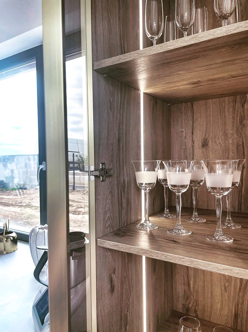 Drewniany regał z podświetleniem LED, na półkach kryształowe kieliszki z białym wzorem. Widok przez otwarte drzwi na okno i krajobraz za nim.