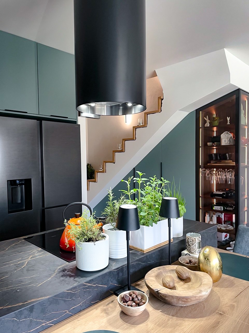 Nowoczesna kuchnia z czarnym okapem cylindrycznym, marmurowym blatem i ziołami w doniczkach. W tle schody i przeszklona witryna z dekoracjami.