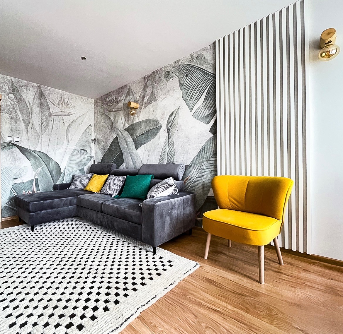 Szary narożnik z kolorowymi poduszkami na tle ściany z motywem liści, żółty fotel i dywan w geometryczne wzory tworzą spójną aranżację wnętrza.