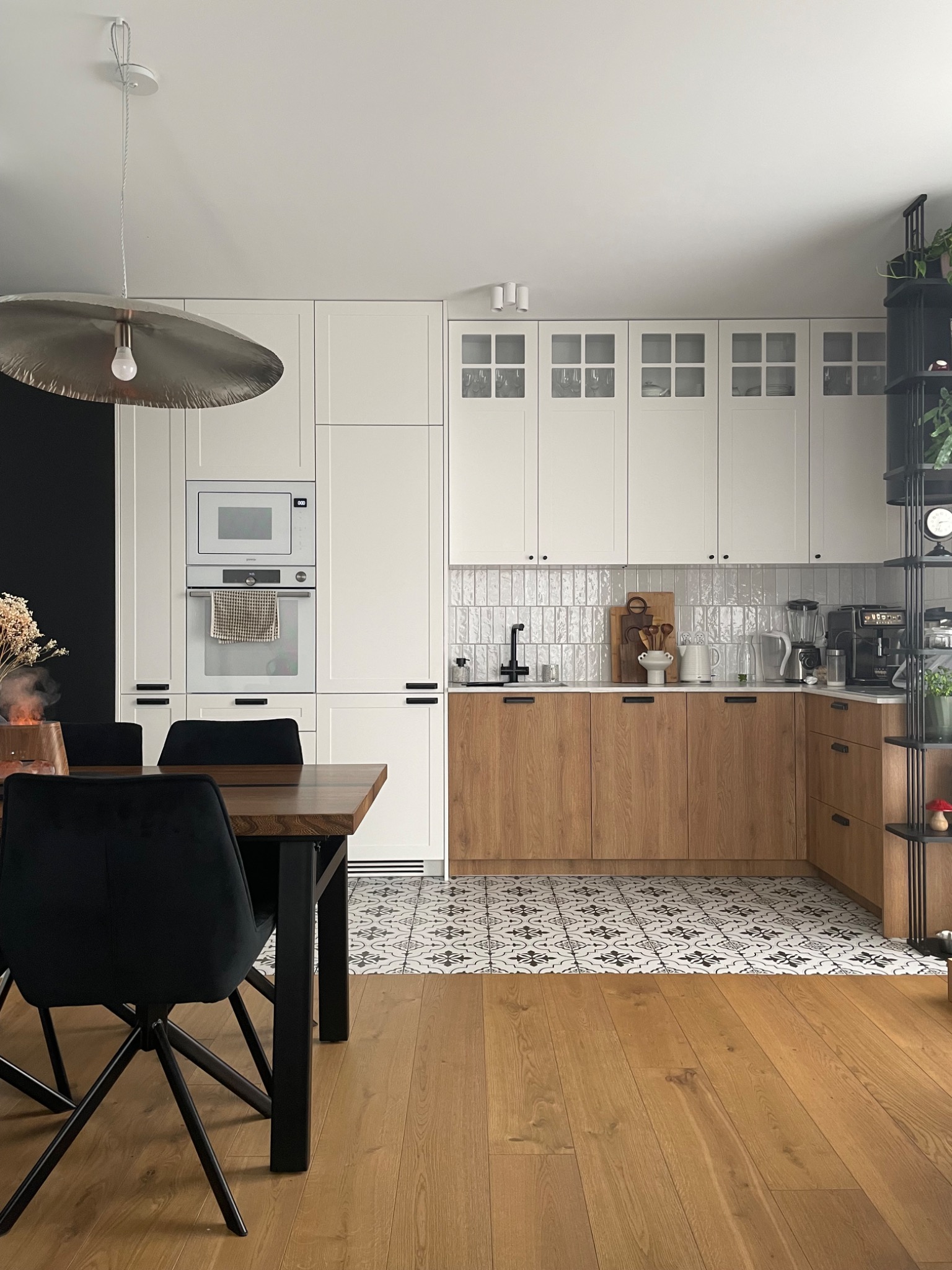 Nowoczesna kuchnia z drewnianymi i białymi szafkami, wzorzystą podłogą i czarnymi akcentami. Widoczny stół z krzesłami i dekoracje.