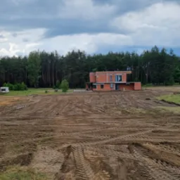 Surowy stan domu z czerwonej cegły w trakcie budowy, otoczony przygotowanym terenem pod budowę, z lasem w tle i pochmurnym niebem.