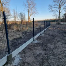 Nowoczesne ogrodzenie panelowe z podmurówką betonową, na tle jesiennego krajobrazu. Widok z perspektywy gruntu, błękitne niebo w tle.