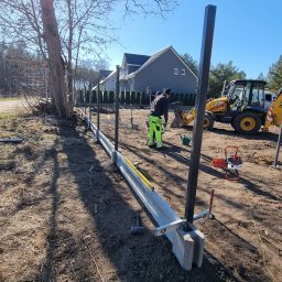 JP-KOP - Montaż ogrodzenia: pracownik w odblaskowej odzieży, czarne słupki ogrodzeniowe w betonowych podstawach, poziomica, wykop, traktor JCB w tle, słoneczny dzień.