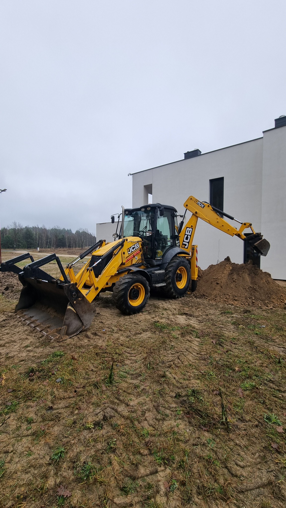 Żółta koparko-ładowarka JCB w trakcie prac ziemnych przy nowoczesnym, białym budynku. Widoczne ślady opon na ziemi i pryzma ziemi. Pochmurne niebo w tle.