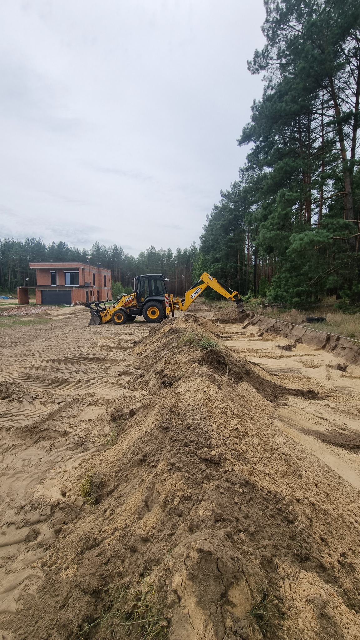 Żółta koparko-ładowarka JCB w Wołowych Lasach podczas prac ziemnych. Wykop pod fundamenty, w tle surowy budynek z czerwonej cegły i las.