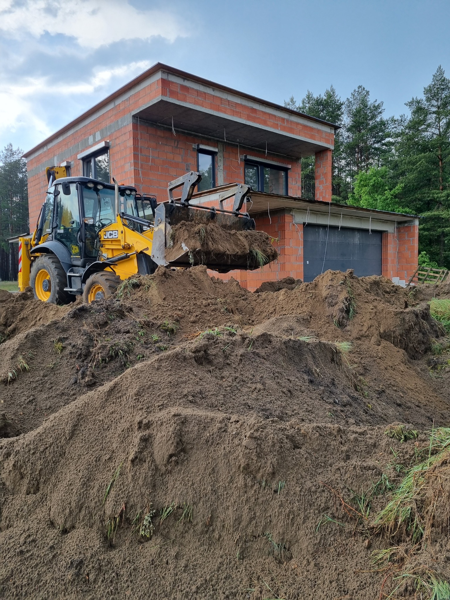 Żółta koparko-ładowarka JCB w trakcie prac ziemnych przy budowie domu z czerwonej cegły. Widoczne hałdy ziemi i fragmenty zieleni.