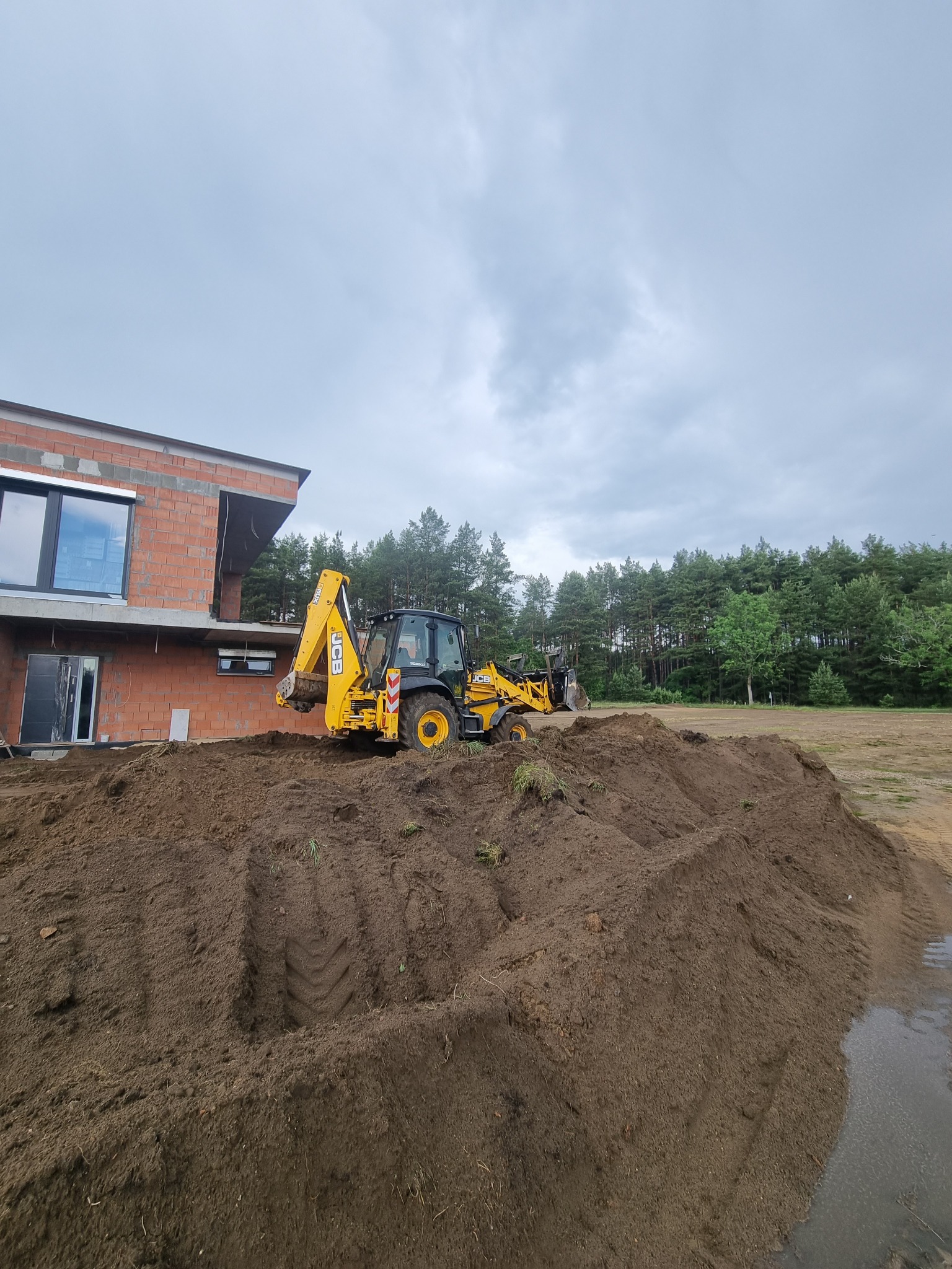 Żółta koparko-ładowarka JCB na stercie ziemi przy budowie domu z czerwonej cegły. W tle drzewa i pochmurne niebo.