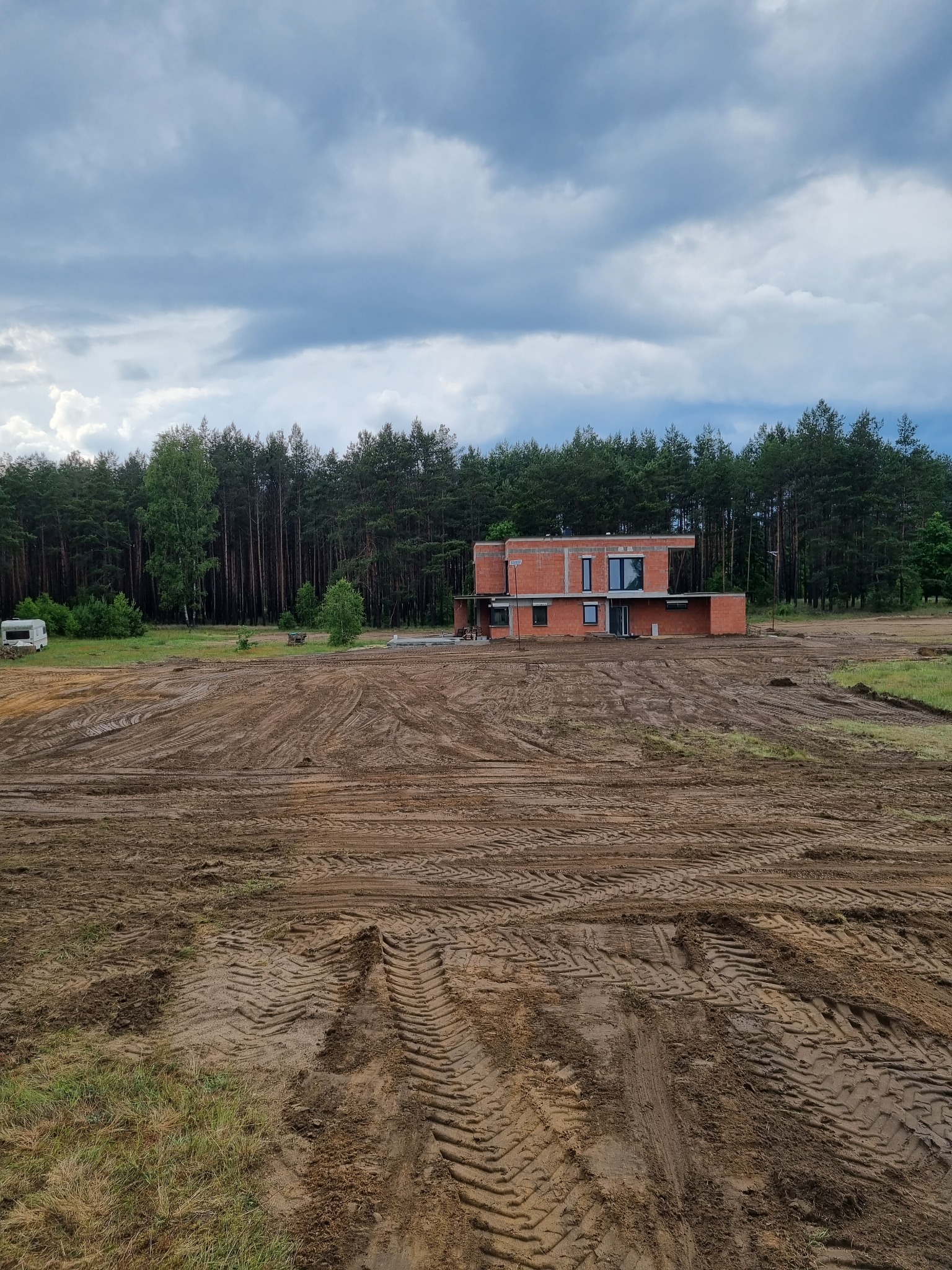 Surowy stan domu z czerwonej cegły w trakcie budowy, otoczony przygotowanym terenem pod budowę, z lasem w tle i pochmurnym niebem.