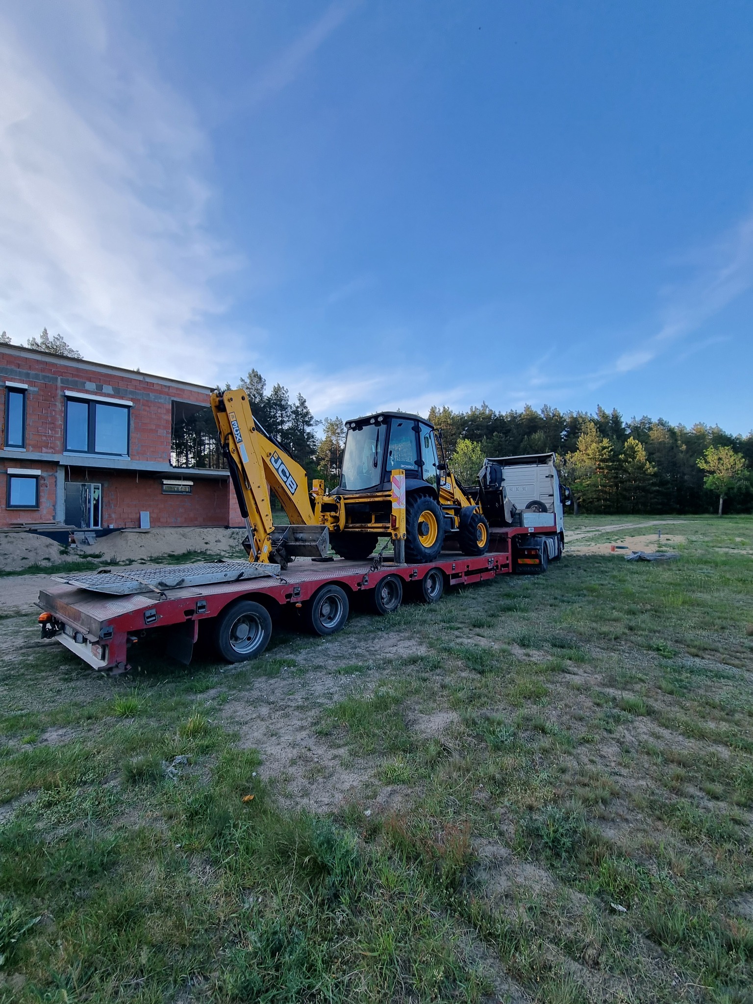 Żółta koparko-ładowarka JCB na niskopodwoziowej platformie transportowej, na tle budynku w trakcie budowy i drzew. Sprzęt gotowy do pracy na placu budowy.