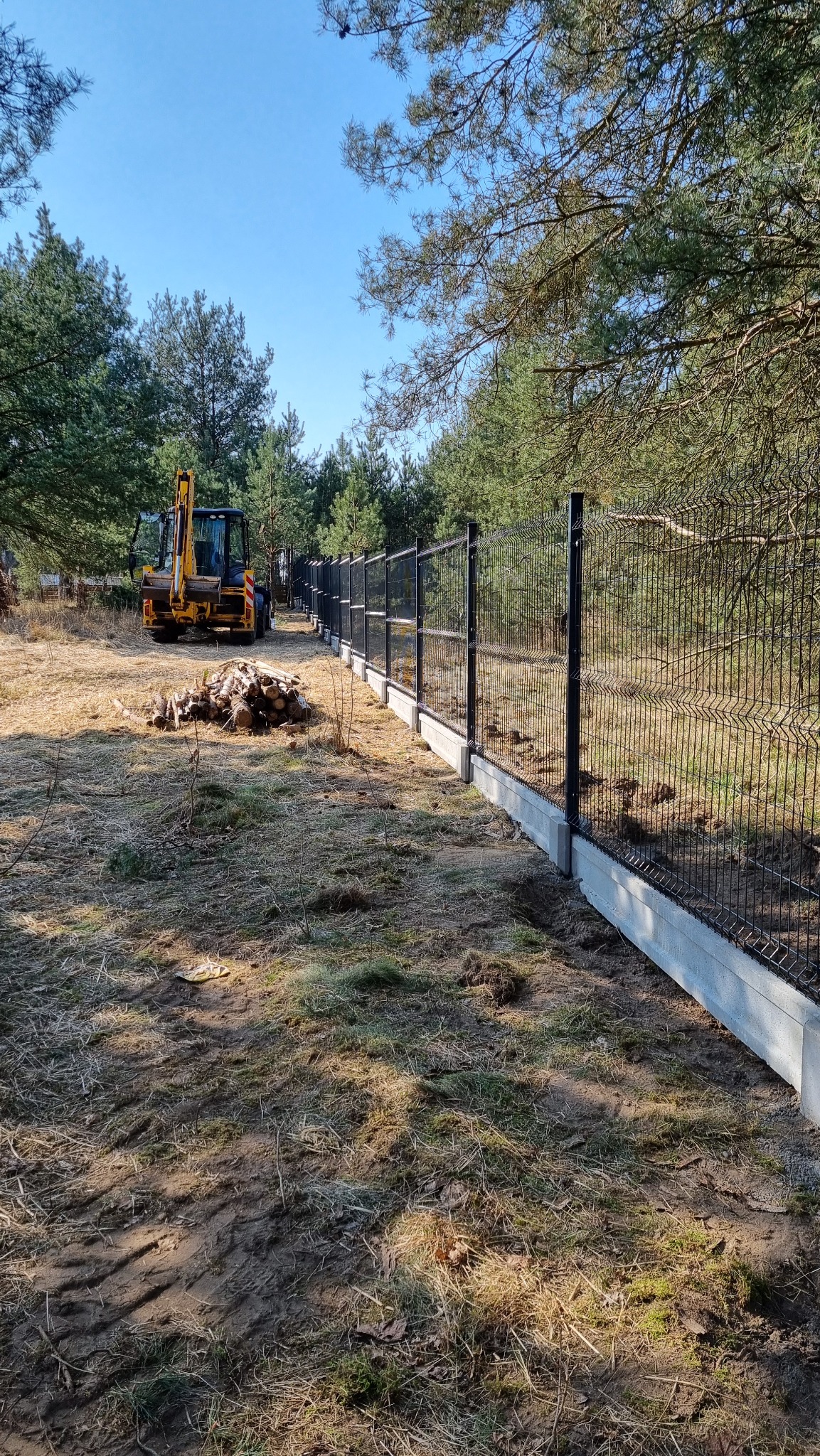 Nowoczesne ogrodzenie panelowe na betonowej podmurówce, z żółtą koparką w tle na leśnej działce. Widok z poziomu gruntu, słoneczny dzień.
