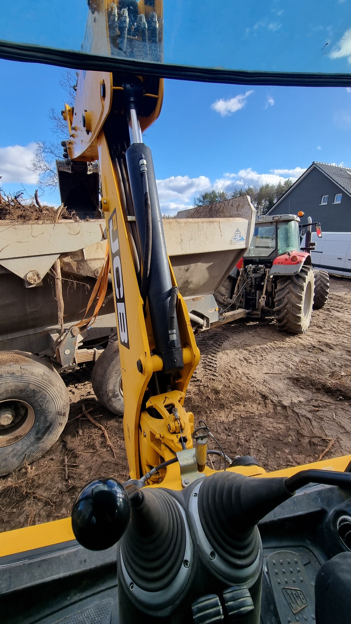Widok z kabiny koparki JCB na przyczepę z ziemią i ciągnik na placu budowy, przy słonecznej pogodzie. Żółte ramię koparki z czarnym siłownikiem.