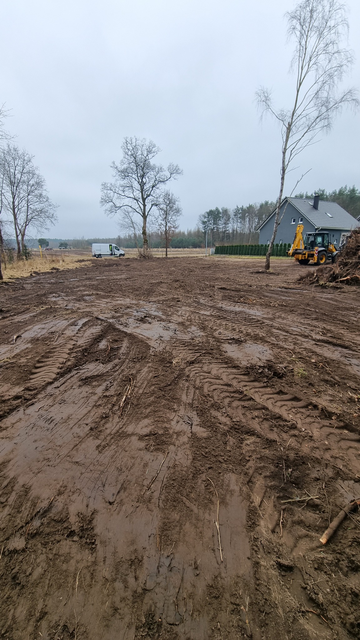 Przygotowany teren pod budowę z widocznymi śladami opon, w tle dom jednorodzinny, drzewa bez liści i pojazd budowlany. Pochmurny dzień.