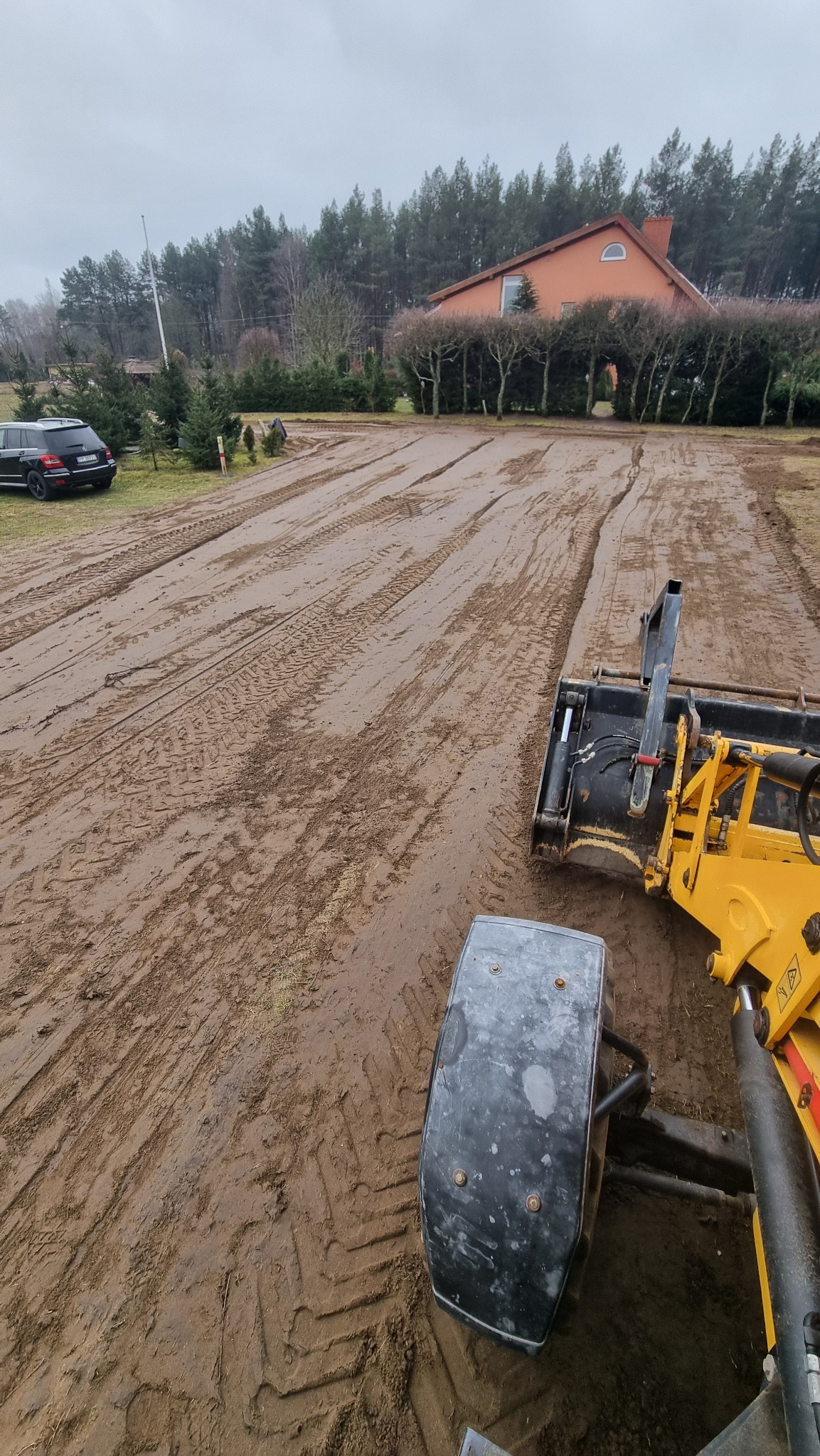 Żółta koparka na placu budowy, przygotowany teren pod inwestycję, ślady opon na błotnistym podłożu, w tle dom i drzewa. Widok z kabiny operatora.