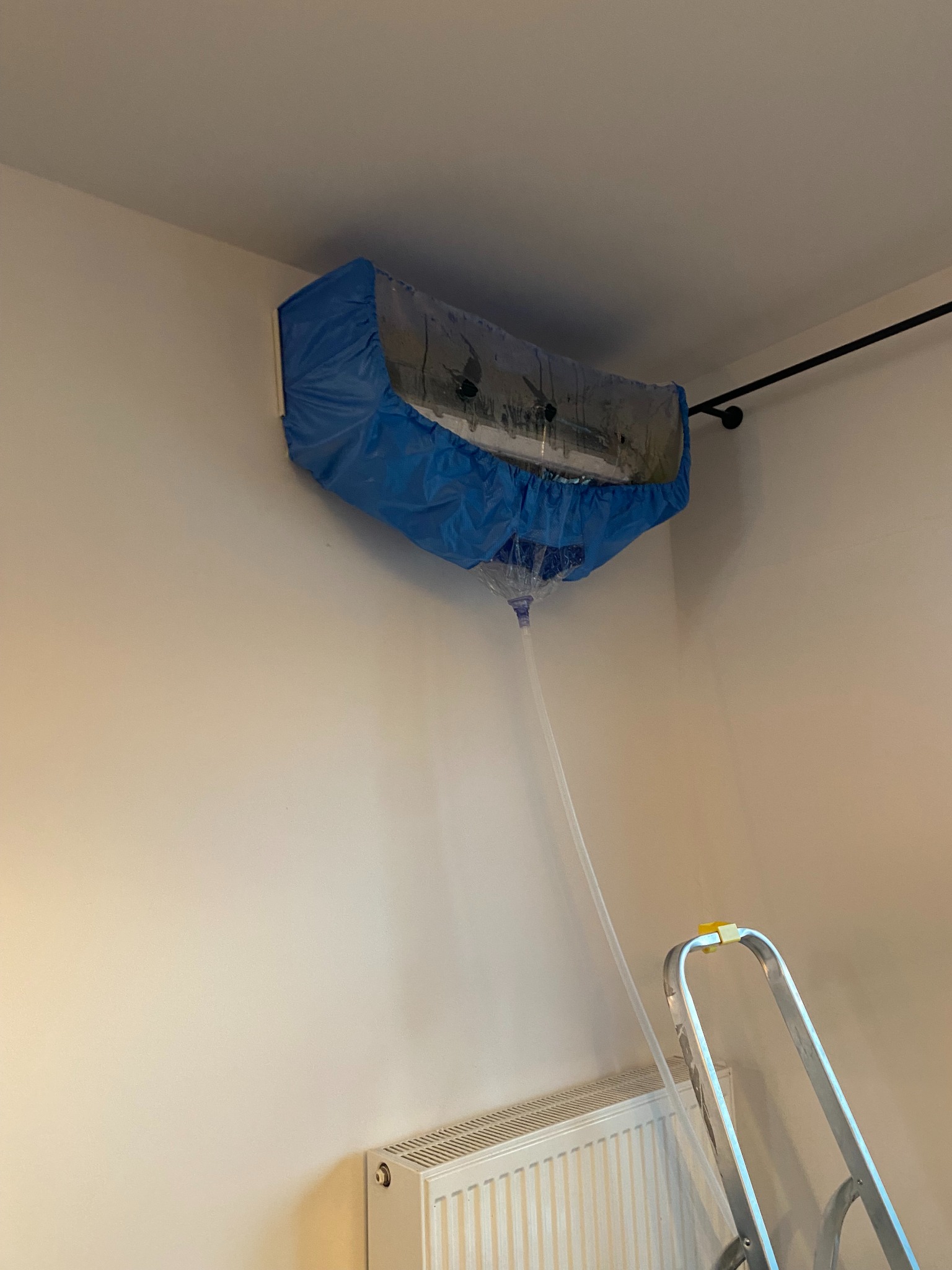 Czyszczenie klimatyzacji naściennej z użyciem specjalnego pokrowca i węża odprowadzającego wodę. Widoczna drabina i grzejnik.