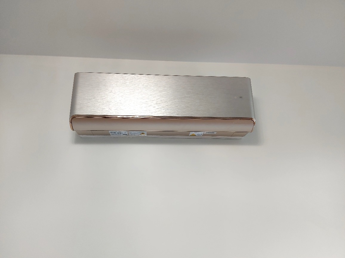 Nowoczesny klimatyzator ścienny w kolorze szczotkowanego srebra z miedzianymi akcentami, zamontowany na białej ścianie. Widok z dołu, oświetlenie dzienne.