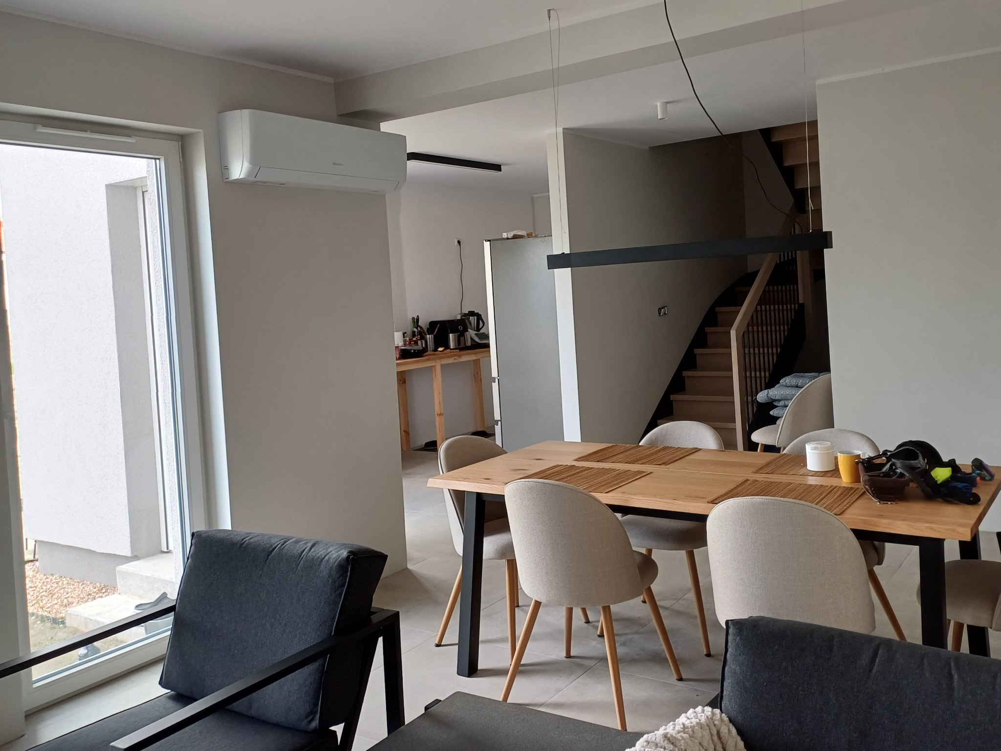 Nowoczesny salon z klimatyzatorem ściennym. Widoczny stół z krzesłami, schody i okno. Minimalistyczny design wnętrza z przewagą bieli i szarości.