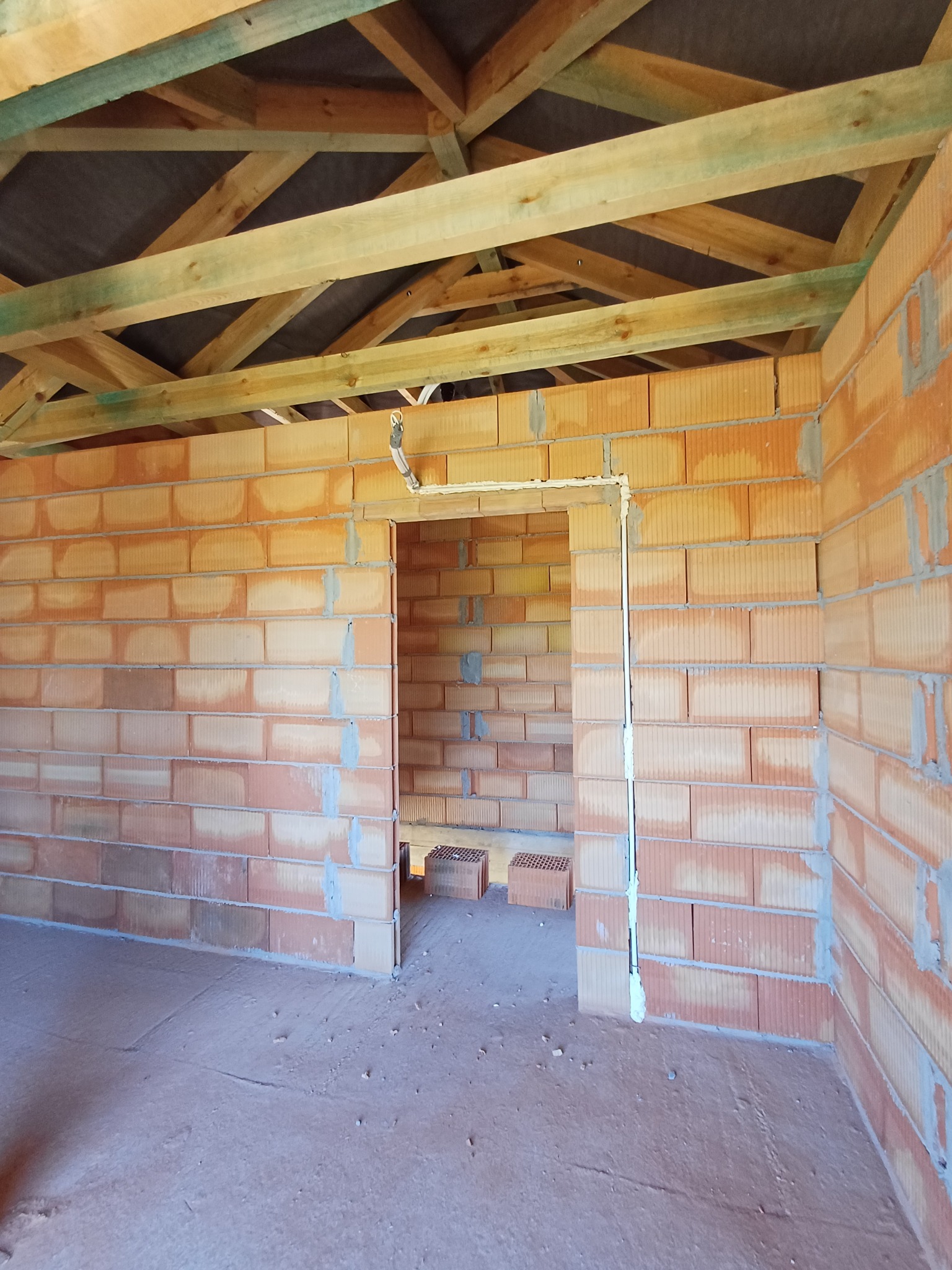 Surowy stan budynku w trakcie budowy: ściany z cegły dziurawki, drewniana konstrukcja dachu, widoczne przewody instalacji elektrycznej przy otworze drzwiowym.