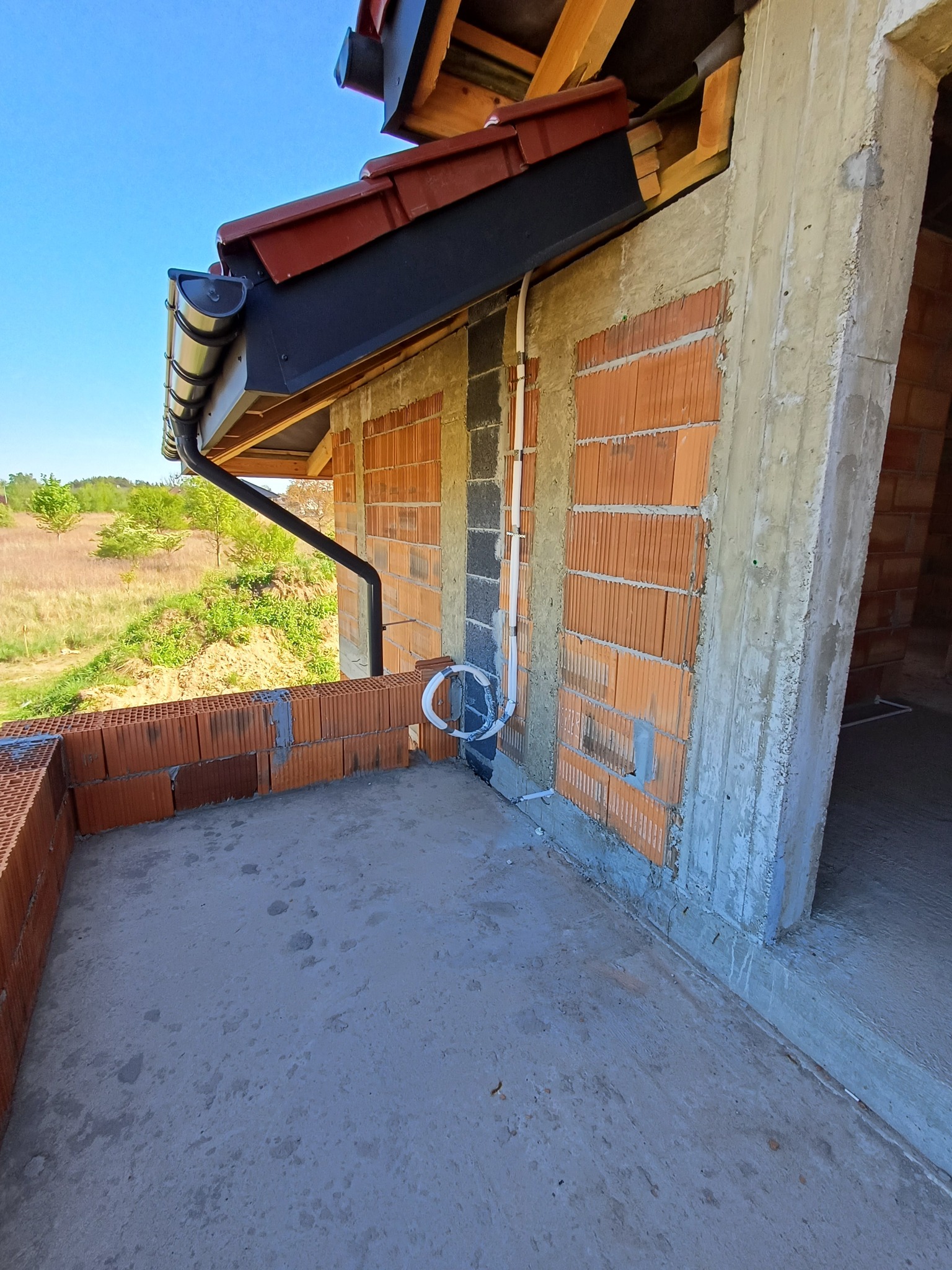 Surowy stan balkonu z ceglaną balustradą i widoczną instalacją elektryczną na ścianie budynku w trakcie budowy. W tle zielony krajobraz.