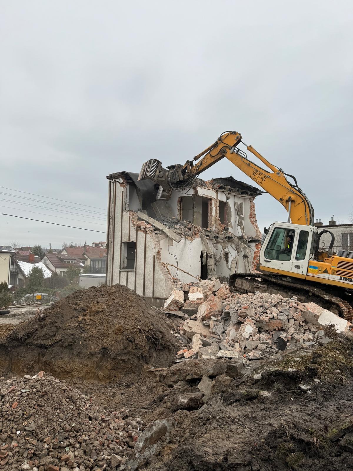 Żółta koparka z operatorem w kabinie, w trakcie rozbiórki budynku z białej cegły, z gruzem i stertą ziemi na pierwszym planie. Widok na zabudowę w tle.