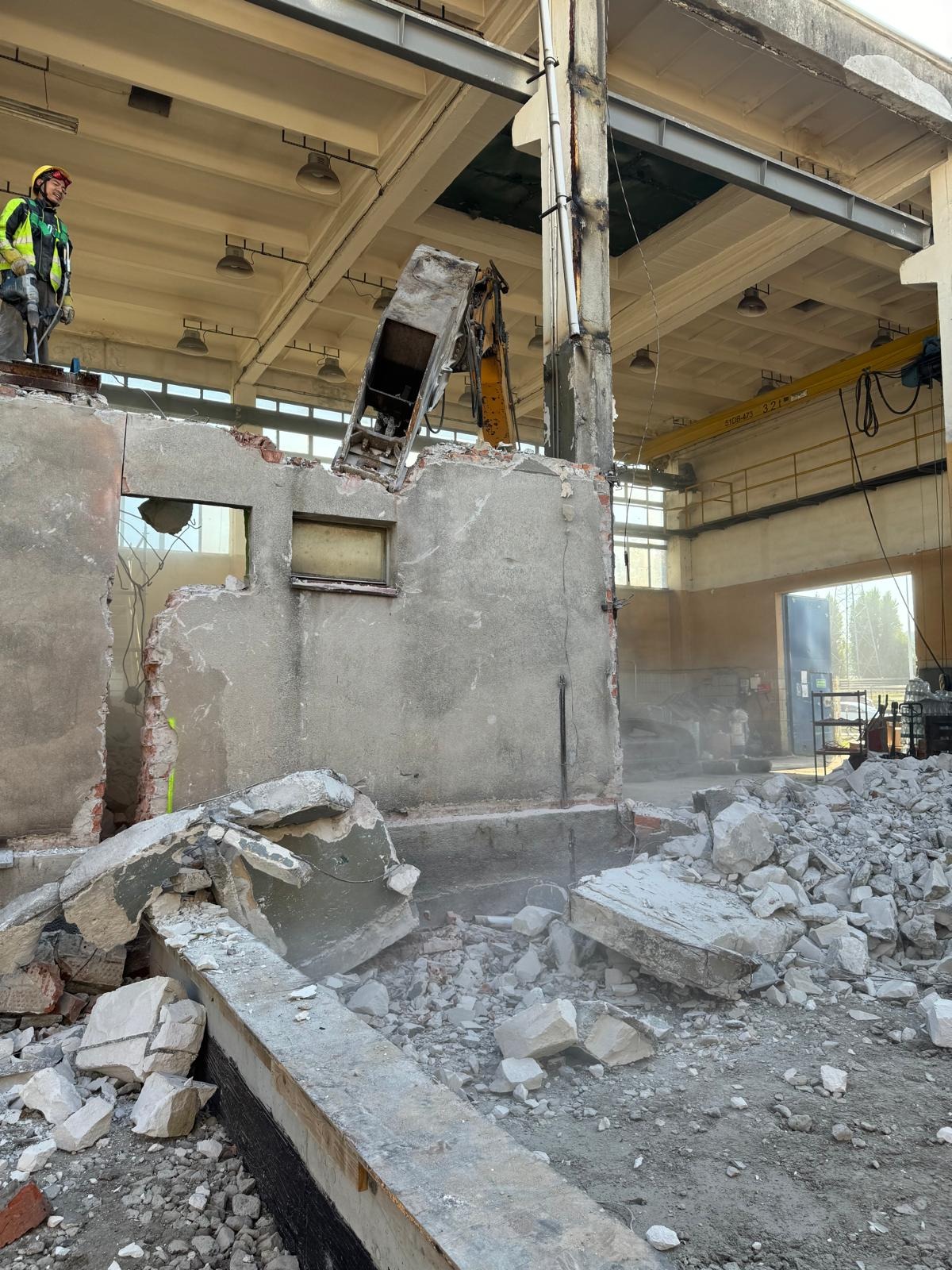 Rozbiórka budynku w hali: operator w kasku, maszyna z wysięgnikiem, gruzowisko, fragmenty ścian z oknami, stalowe konstrukcje stropu i suwnica.