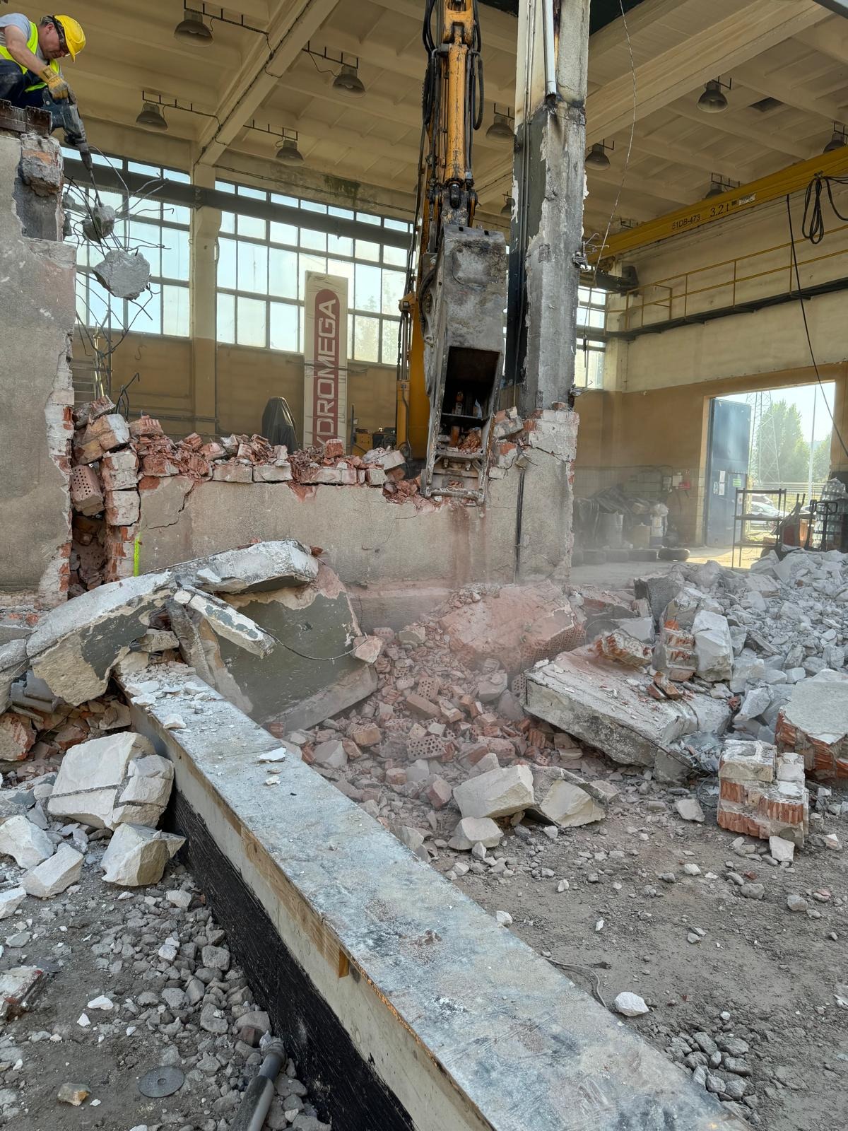 Demontaż ściany w hali: operator koparki z młotem hydraulicznym kruszy mur, widoczne cegły, beton, gruz. Robotnik w kasku nadzoruje prace. W tle konstrukcja hali.