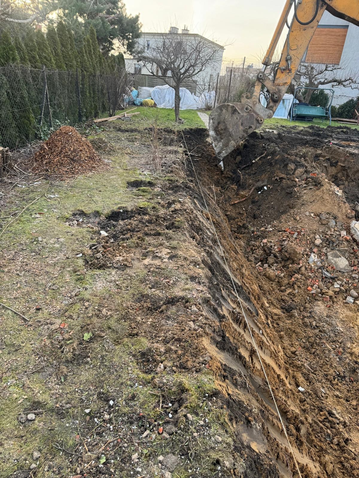 Żółta koparka w trakcie wykonywania wykopu w ziemi, widoczna linia wyznaczająca trasę, sterta liści i budynek w tle. Prace ziemne w ogrodzie.