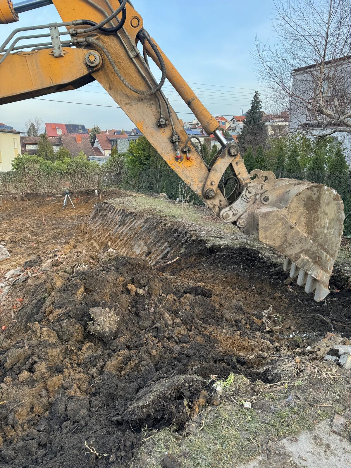 Żółta koparka w trakcie prac ziemnych na działce, widoczny wykop i sterta ziemi. W tle zabudowa jednorodzinna i zieleń. Na drugim planie niwelator.