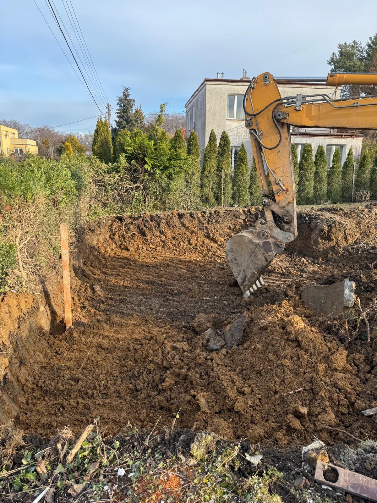 Żółta koparka w trakcie wykopu pod fundamenty na działce z zielenią. Widoczne ślady pracy maszyny w ziemi, budynek w tle. Słoneczny dzień.