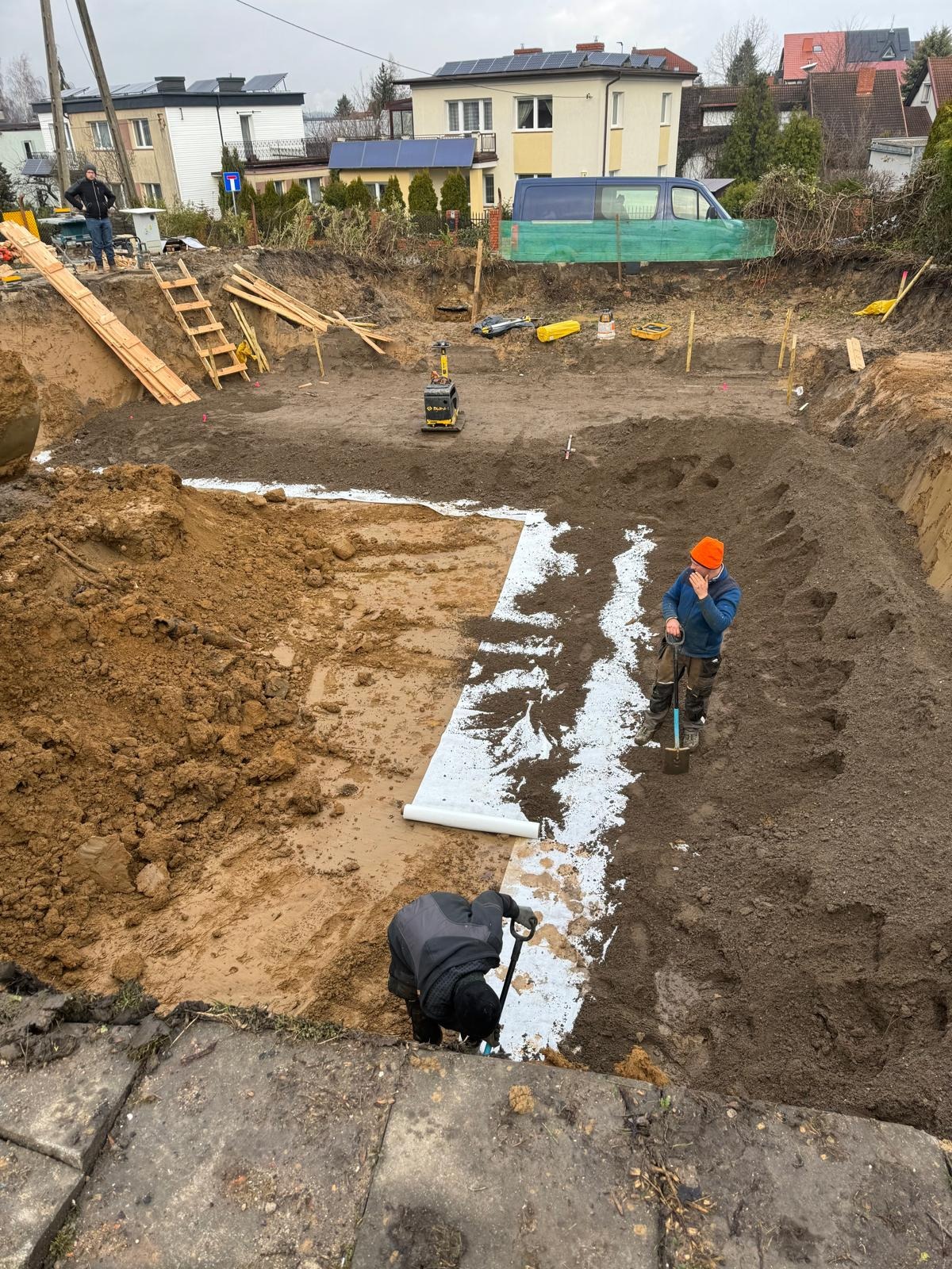 Prace ziemne: dwóch robotników rozkłada białą geowłókninę w wykopie pod budowę. W tle widoczny sprzęt budowlany, drewniane elementy konstrukcyjne oraz zabudowa mieszkalna.
