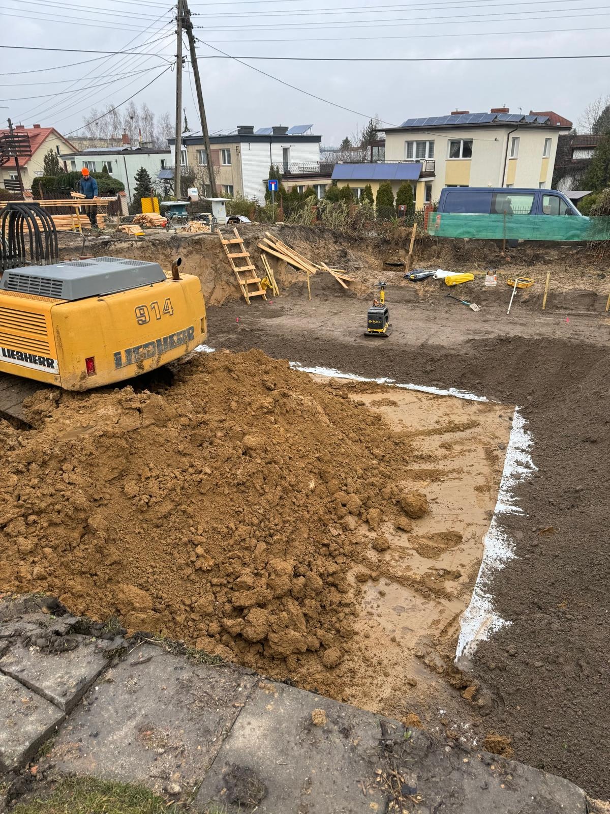 Żółta koparka Liebherr 914 przy wykopie fundamentów, widoczne deski, drabina i ubijarka. Na pierwszym planie hałda ziemi i przygotowane podłoże.