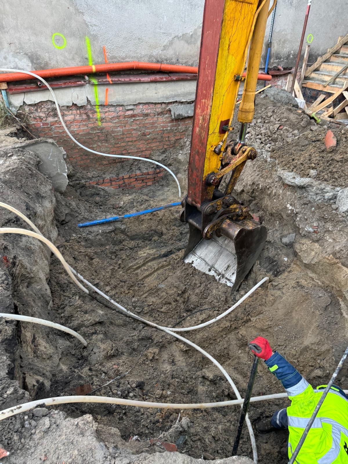 Żółta koparka w wykopie z widocznymi rurami i instalacjami w Gdyni. Prace ziemne w toku, widoczny fragment muru z cegły i pracownik w odblaskowej kamizelce.