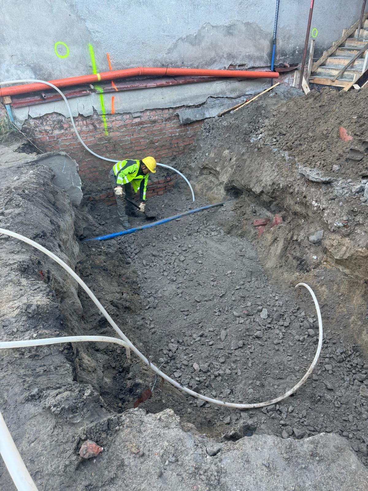 Pracownik w żółtym kasku kopie dół obok fundamentów budynku. Widoczne rury instalacyjne i oznaczenia na ścianie. Etap prac ziemnych.
