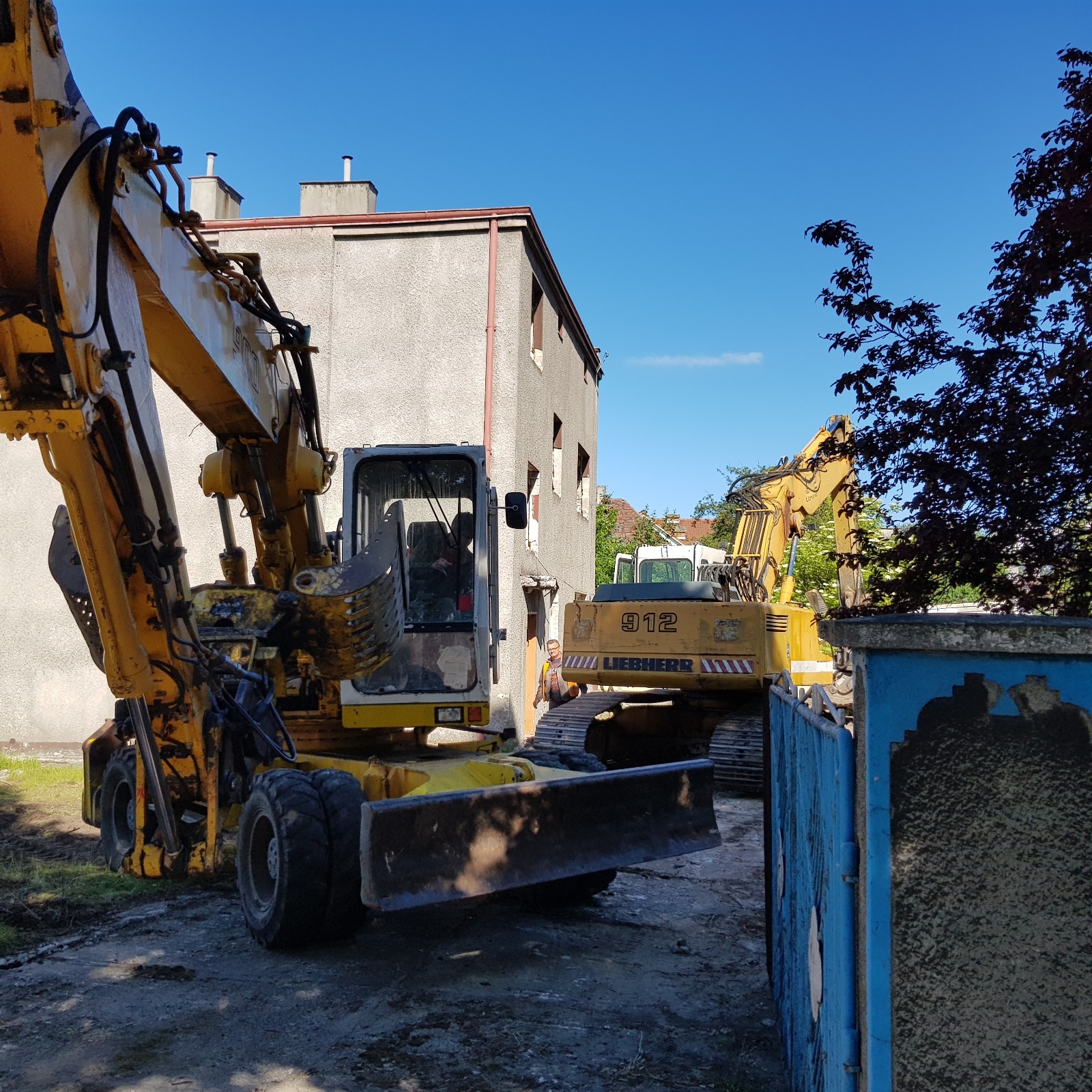 Żółte koparki Liebherr na placu budowy w Gdyni, na tle budynku z wyburzonymi oknami. Widoczny operator maszyny.