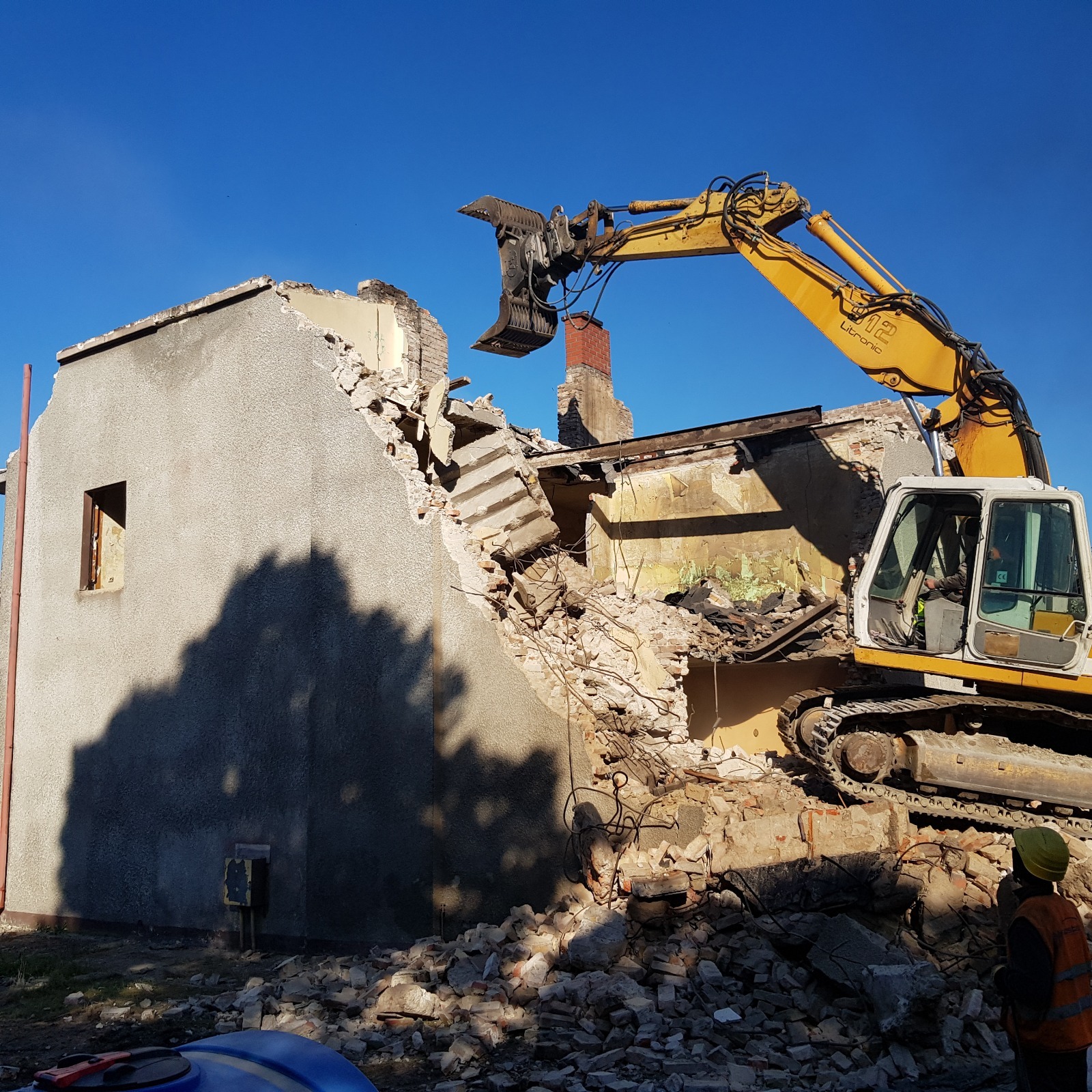 Żółta koparka Litronic burzy budynek z cegły i betonu na tle błękitnego nieba. W tle widoczny komin. Na pierwszym planie gruz i fragment sylwetki osoby w kamizelce.