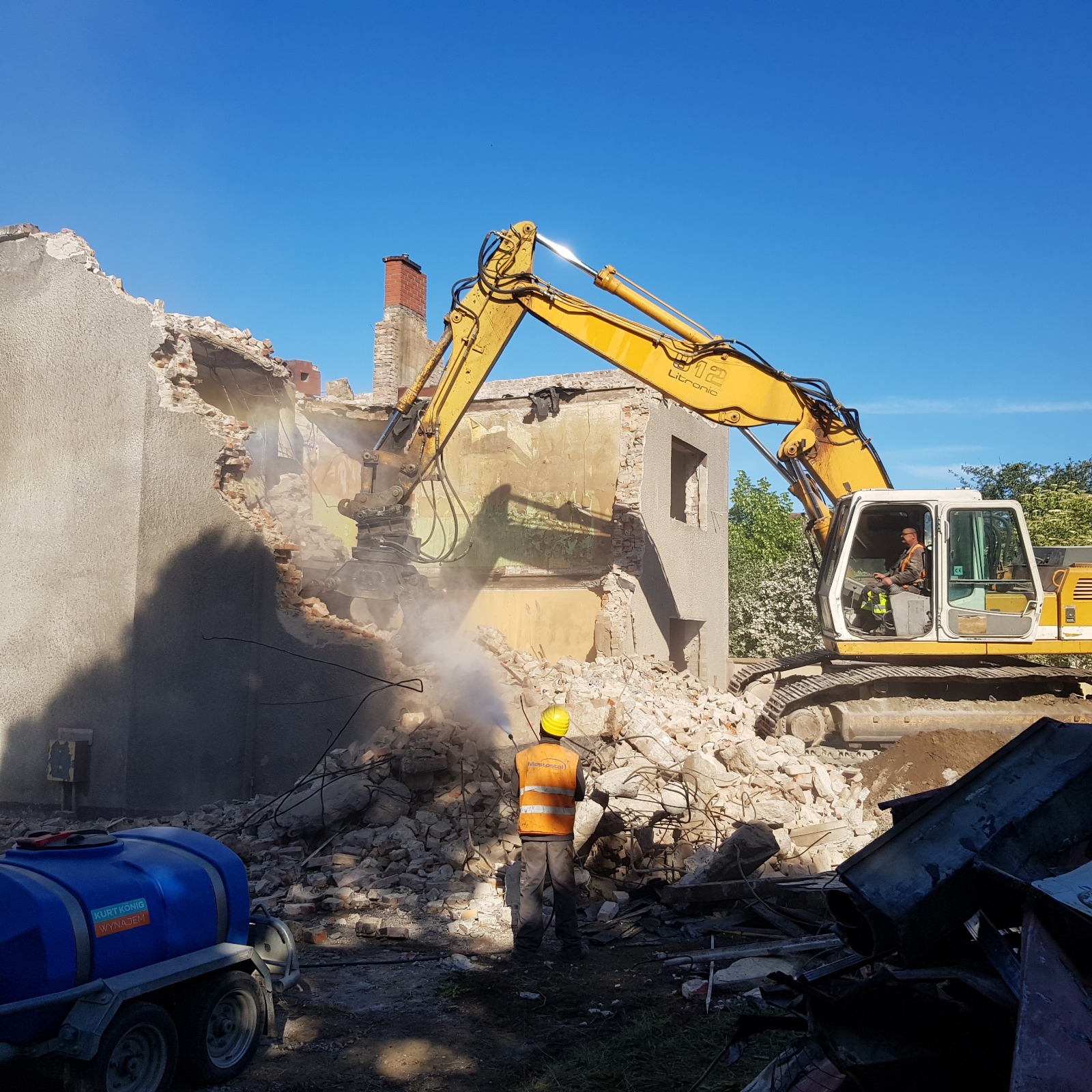 Żółty koparka z hydraulicznym młotem rozbiera budynek, w tle pracownik spryskuje gruz wodą, by ograniczyć zapylenie. Widoczne pozostałości ścian i gruzowisko.