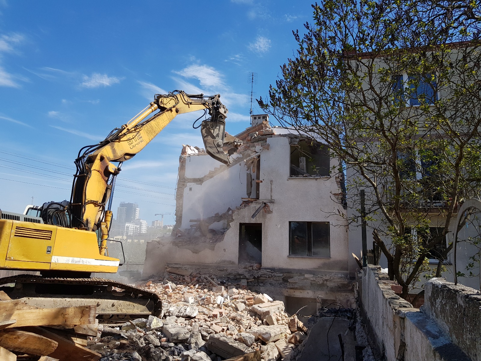 Żółta koparka z chwytakiem burzy dwupiętrowy budynek z białej cegły na tle błękitnego nieba. W tle panorama miasta i drzewo z zielonymi liśćmi.