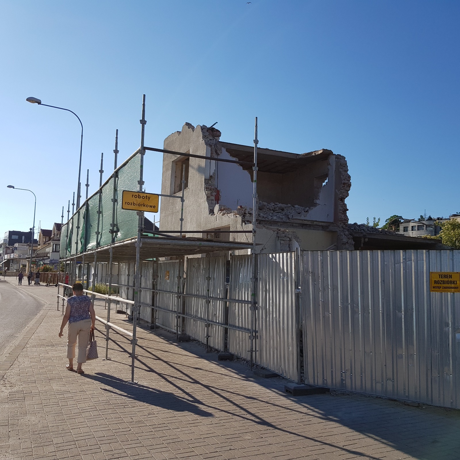 Rozbiórka budynku w trakcie. Widoczne rusztowania, zabezpieczenia, tablice informacyjne. Kobieta przechodzi obok. Częściowo zburzona konstrukcja.